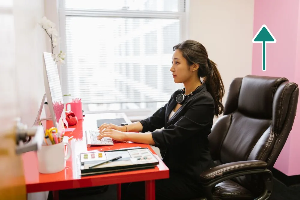 Side view of a woman working at a desk with the top of the monitor at about eye level, sitting back in an adjustable office chair, with an arrow above the chair pointing up